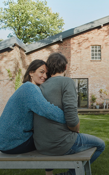 Ein Mann und eine Frau sitzen mit dem Rücken zur Kamera vor einem Haus. Die Frau umarmt den Mann von hinten. Sie blickt über die Schulter in die Kamera.