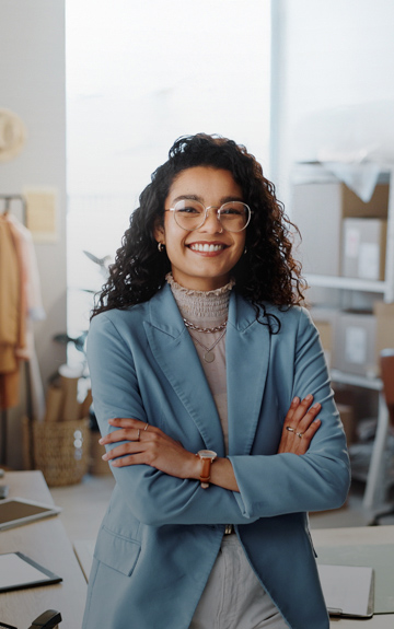Eine Schneiderin steht in ihrer Werkstatt. Sie hat die Arme verschränkt und blickt in die Kamera.