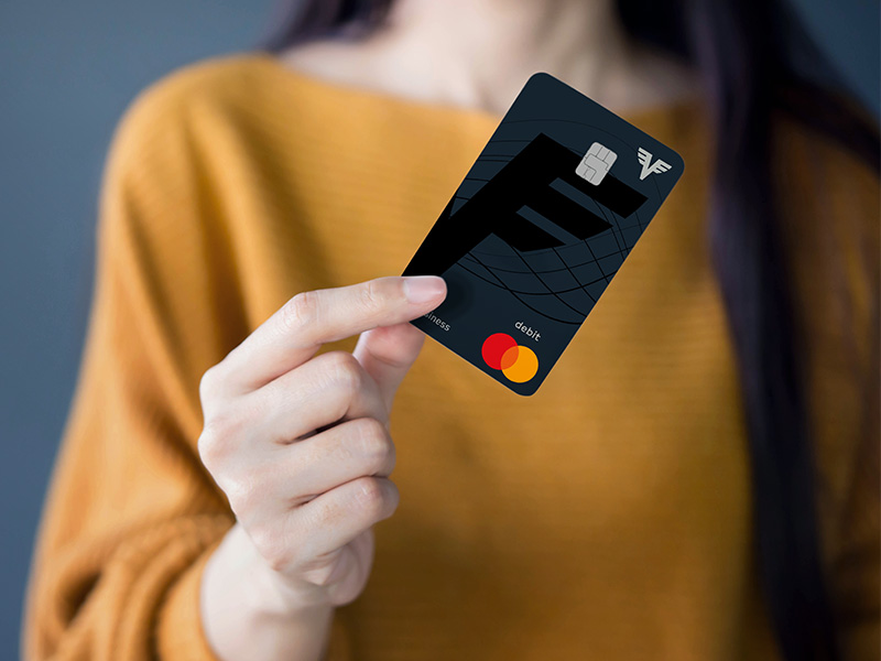 Eine Frau in einem gelben Pullover hält die Volksbank Debit Mastercard Business in der Hand. Man kann ihr Gesicht nicht sehen.