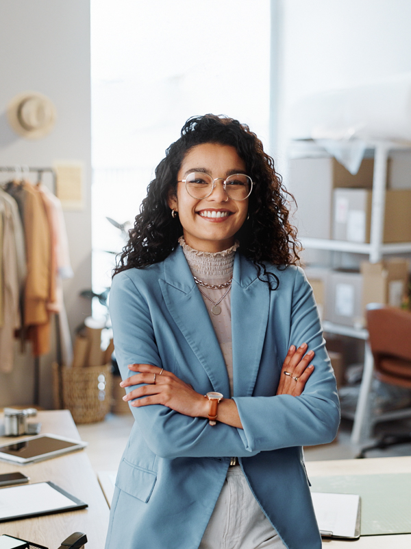 Eine junge Schneiderin steht in ihrer Schneiderei. Sie hat die Arme verschränkt und blickt selbstbewusst in die Kamera.