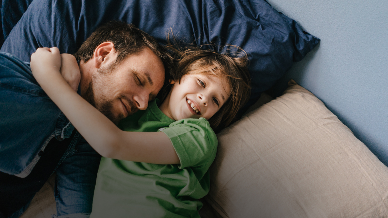 Ein Vater und sein Sohn liegen auf einer Couch. Sie umarmen sich und lächeln.