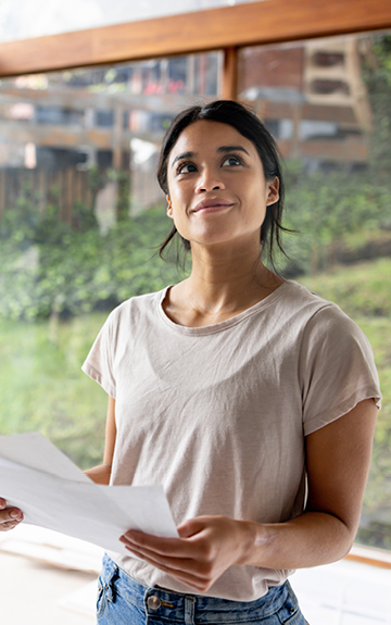 Eine Frau steht vor einer Fensterfassade, hält einen Plan in der Hand und lächelt