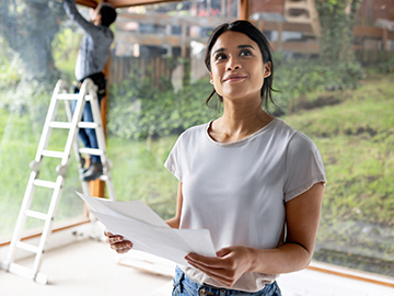 Eine junge Frau steht in ihrem neuen Haus, das gerade gebaut wird. Sie hat einen Bauplan in der Hand.